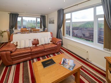 A living room with a leather sofa and wooden coffee table next to large windows at Fenny Marina Cottage in Fenny Bentley