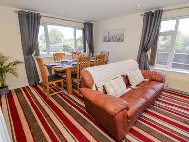 A dining area with wooden chairs and table next to a leather sofa in a room with windows and striped carpet at Fenny Marina Cottage in Fenny Bentley