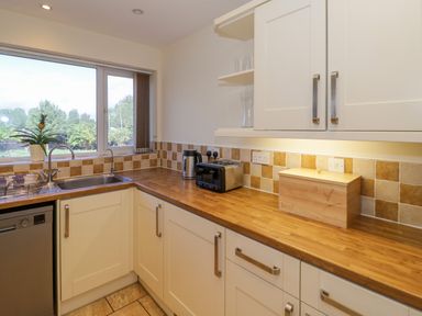 A kitchen with white cabinets wooden countertops a sink toaster kettle wooden bread box and a window at Fenny Marina Cottage in Fenny Bentley