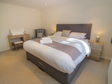 A bedroom with a large bed two bedside tables a dressing table and a plaid armchair at Fenny Marina Cottage in Fenny Bentley