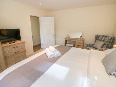 A bedroom with a bed folded towels a chest of drawers a television a dressing table and a checked armchair at Fenny Marina Cottage in Fenny Bentley