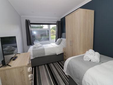 A bedroom with two single beds a wooden wardrobe a chest of drawers and a window with dark curtains at Fenny Marina Cottage in Fenny Bentley