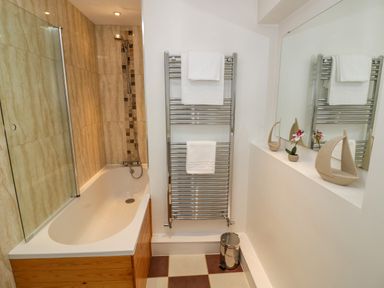 A bathroom with a bathtub white towels on a heated towel rail a small trash bin and decorative boat and flower on a shelf at Fenny Marina Cottage in Fenny Bentley