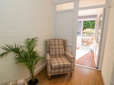 A hallway with a plaid armchair and a potted plant leading to a sunroom with wooden chairs at Fenny Marina Cottage in Fenny Bentley
