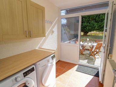 A laundry room with washing machine and dryer under a wooden countertop and a door leading to an outdoor wooden deck with table and chairs at Fenny Marina Cottage in Fenny Bentley