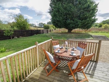 A wooden deck with a table and four chairs with cushions and cups in a fenced garden at Fenny Marina Cottage in Fenny Bentley