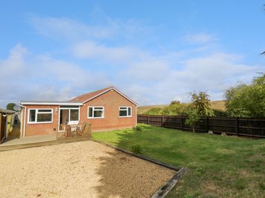 A brick house with a small wooden deck and green lawn with a gravel area and wooden fence in the garden at Fenny Marina Cottage in Fenny Bentley