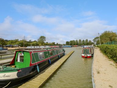 Canal boats docked along wooden piers with a gravel path and cars parked nearby at Fenny Marina Cottage in Fenny Bentley