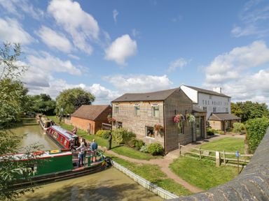 Canal boats on water near buildings including The Wharf Inn with people standing outside on grass at Fenny Marina Cottage in Fenny Bentley