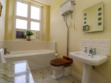 A bathroom with a bathtub a toilet with wooden seat a pedestal sink and a window with flowers at North Wing Middleton Hall Estate near Belford