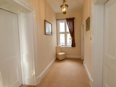 A hallway with beige carpet cream walls a window with a brown curtain a cream chair two framed pictures and white doors at North Wing in Middleton Hall Estate near Belford