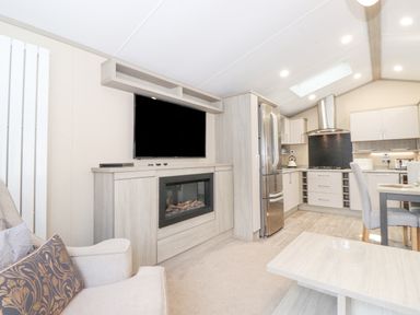 An open living area with a wall-mounted television above an electric fireplace a beige armchair a stainless steel refrigerator a kitchen with cabinets and a dining table with chairs at Tawny Owl Lodge Coniston 4 White Cross Bay near Windermere