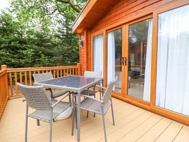 An outdoor wooden deck with a glass-top table and four wicker chairs outside a cabin with sliding glass doors at Tawny Owl Lodge in Coniston 4 White Cross Bay near Windermere