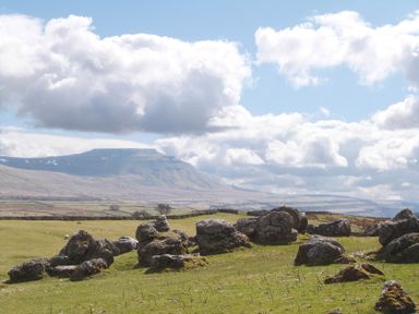 A grassy field with large rocks scattered and mountains in the background under a partly cloudy sky at Lapwing in Chapel Le Dale near Ingleton
