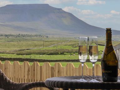 A view of a mountain and green fields with sheep behind a wooden fence with a table holding two champagne glasses and a bottle at Lapwing in Chapel Le Dale near Ingleton
