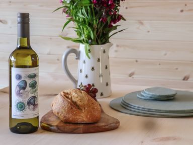 A table with a bottle of wine, a loaf of bread on a cutting board, grapes, a jug with flowers, and stacked plates at Lapwing in Chapel Le Dale near Ingleton