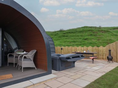 An outdoor patio area with two wicker chairs and a small table under a curved wooden structure a picnic table with items on it a closed hot tub a chiminea and a wooden fence with grassy hills in the background at Lapwing in Chapel Le Dale near Ingleton
