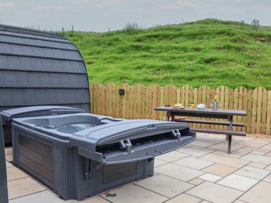 An outdoor patio with a hot tub and a picnic table with drinks and a book by a wooden fence and grassy hill at Lapwing in Chapel Le Dale near Ingleton