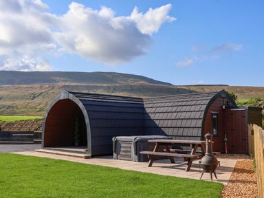 An outdoor area with a curved wooden cabin a picnic table and a chiminea on a paved patio at Lapwing in Chapel Le Dale near Ingleton