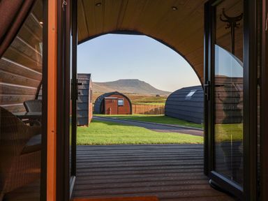 View from inside a wooden pod with open glass doors showing patio chairs lawn path and other pods with a mountain in the background at Oyster Catcher in Chapel Le Dale near Ingleton