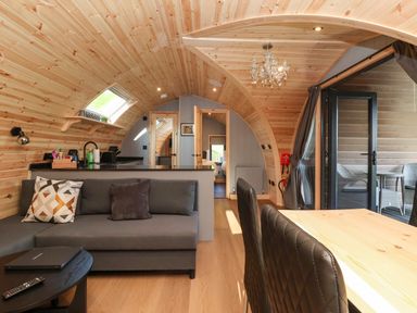A curved wooden interior with a gray sofa and cushions a black coffee table a wooden dining table with black chairs a chandelier and glass doors leading to an outdoor seating area at Oyster Catcher in Chapel Le Dale near Ingleton