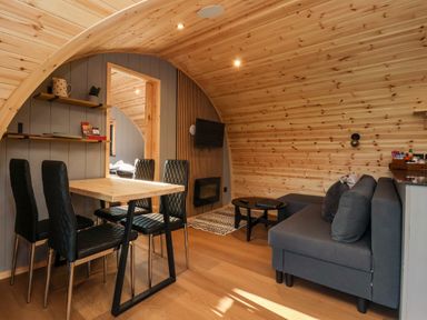 A dining area with a wooden table and four black chairs next to a living area with a gray sofa and a small black table in a wooden cabin interior at Oyster Catcher in Chapel Le Dale near Ingleton