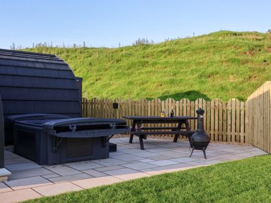 An outdoor patio with a hot tub wooden picnic table and chiminea surrounded by a wooden fence and grassy hill at Oyster Catcher in Chapel Le Dale near Ingleton