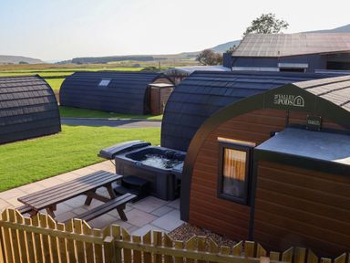 Several wooden pods with curved roofs on a grassy area with a picnic table and hot tub at Lune Valley Pods Chapel Le Dale near Ingleton