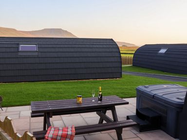 A picnic table with a blanket and bottle on it next to a hot tub outside with two modern curved buildings in the background at Oyster Catcher in Chapel Le Dale near Ingleton
