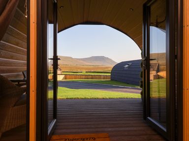 View through open glass doors onto a wooden porch lawn a curved gray structure and a mountain in the distance at Heron in Ingleton