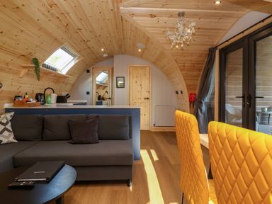 A living area with a gray sofa a black round table yellow quilted chairs and wooden walls at Heron in Ingleton