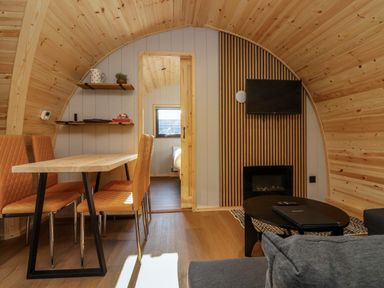 A living room with wooden walls and ceiling featuring a dining table with four chairs a black coffee table a television mounted above a fireplace and a doorway leading to a bedroom at Heron in Ingleton