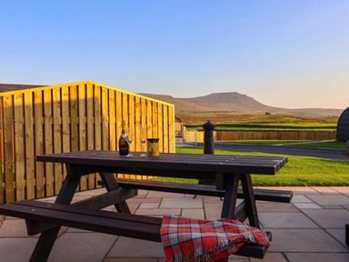 An outdoor wooden picnic table with drinks glasses bottle and a blanket on a patio with a wooden fence and hills in the background at Heron in Ingleton