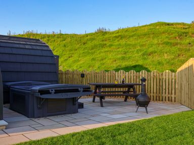An outdoor patio with a hot tub a wooden picnic table with bottles and glasses and a metal fire pit at Heron in Ingleton