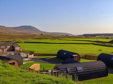 Curved small wooden cottages on green fields with a mountain in the background at Heron in Ingleton