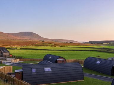 Small curved black roof cabins on green grass fields with a mountain in the background at Heron in Ingleton