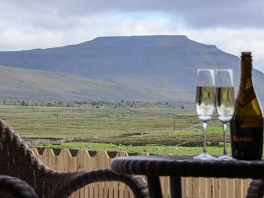 A view of a mountain and green fields with sheep from a balcony with wicker chairs a wooden fence a bottle and two glasses of champagne at Wren in Ingleton