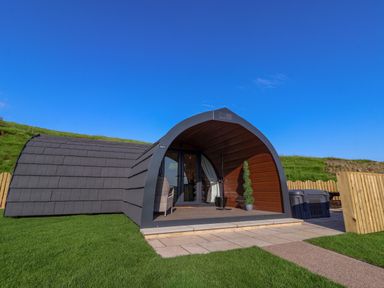 A small modern curved cabin with a patio, chair, and plant on grass at Wren Ingleton