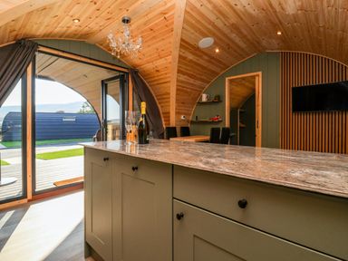 Interior view of a curved wooden room with a kitchen island containing a bottle and glasses and a table and chairs near an open glass door at Wren in Ingleton