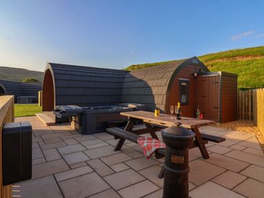 An outdoor patio area with a wooden picnic table, hot tub, curved cabin, and grassy hill in the background at Wren in Ingleton