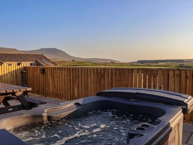 An outdoor area with a hot tub wooden picnic table and a wooden fence with hills in the background at Wren in Ingleton