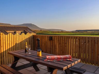 A wooden picnic table with a bottle of wine two glasses a tin and a folded blanket on a fenced patio overlooking green fields and hills at Wren in Ingleton
