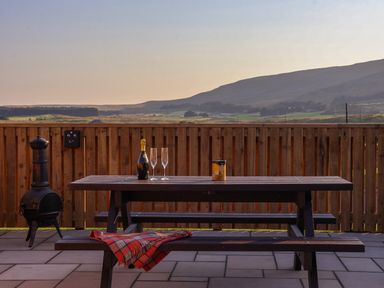 An outdoor wooden picnic table with two glasses and a bottle on top a plaid blanket on the bench and a chiminea next to the fence with a landscape view at Wren in Ingleton