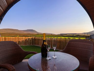 A round glass table with a bottle and two glasses between two wicker chairs on a covered patio overlooking a fenced grassy yard and distant hills at Barn Owl in Chapel Le Dale near Ingleton