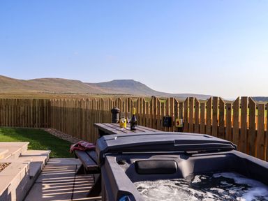An outdoor area with a hot tub a wooden picnic table with drinks and glasses and a wooden fence with hills in the background at Barn Owl in Chapel Le Dale near Ingleton
