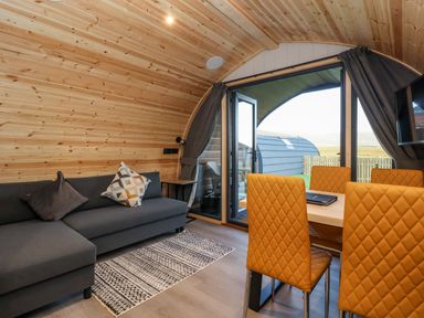 A wooden interior living space with a dark grey sofa and patterned cushions near orange quilted dining chairs around a table at Barn Owl in Chapel Le Dale near Ingleton