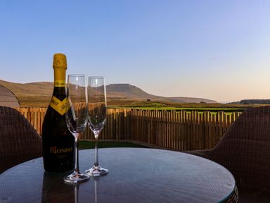 A bottle of sparkling wine with two glasses on a round table outside with a wooden fence and hills in the distance at Barn Owl in Chapel Le Dale near Ingleton