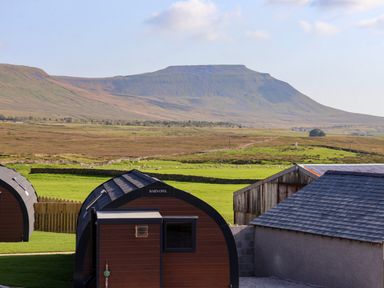 Two curved wooden cabins a wooden fence sheds green fields and a flat topped hill in the background at Barn Owl in Chapel Le Dale near Ingleton
