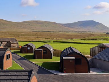 Small group of curved wooden cabins on grassy plots with a paved road and hills in the background at Barn Owl Chapel Le Dale near Ingleton