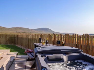 An outdoor area with a hot tub next to a picnic table with drinks and glasses and a wooden fence in the background at Curlew in Chapel Le Dale near Ingleton